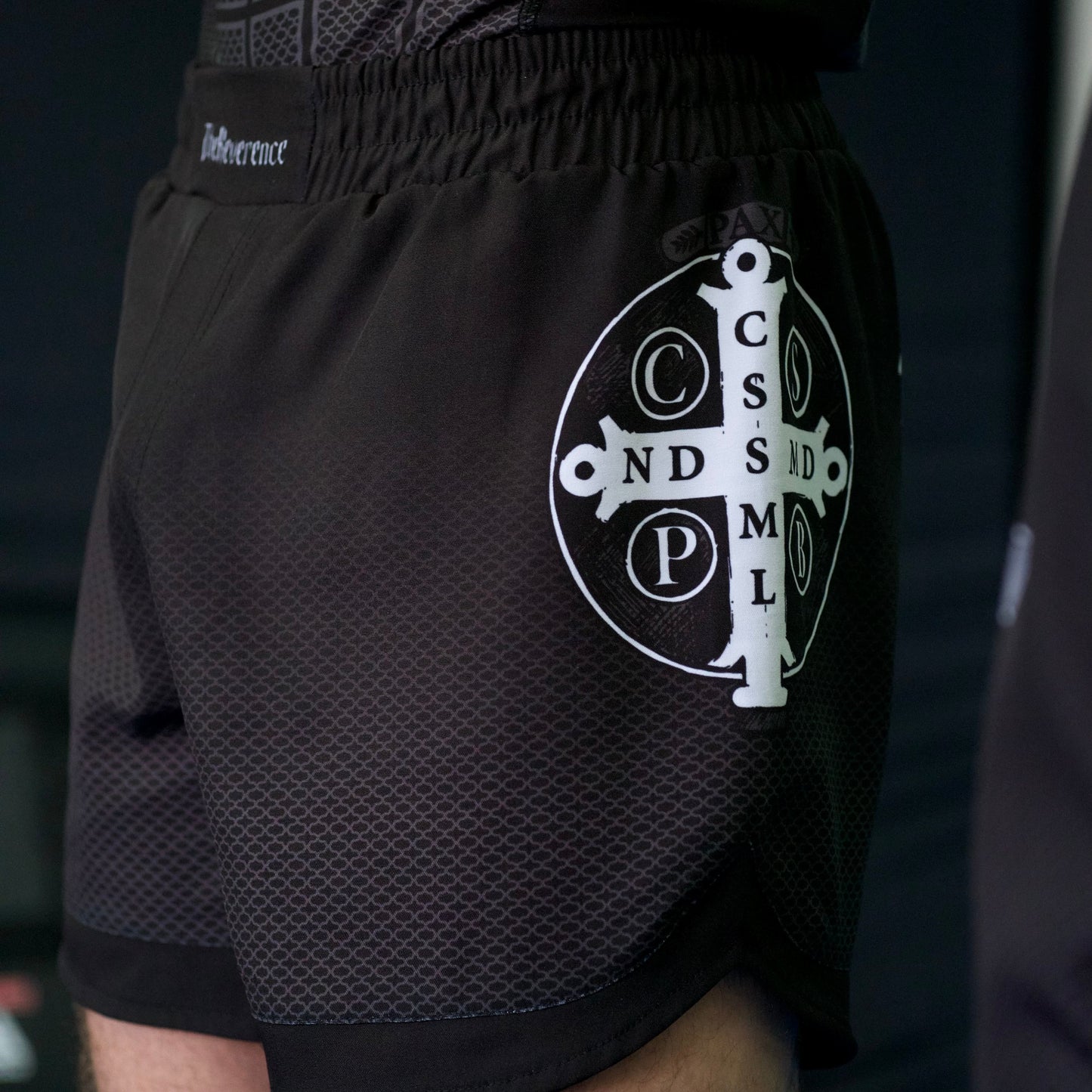 Person wearing black athletic shorts with a logo and text, standing against a dark background.