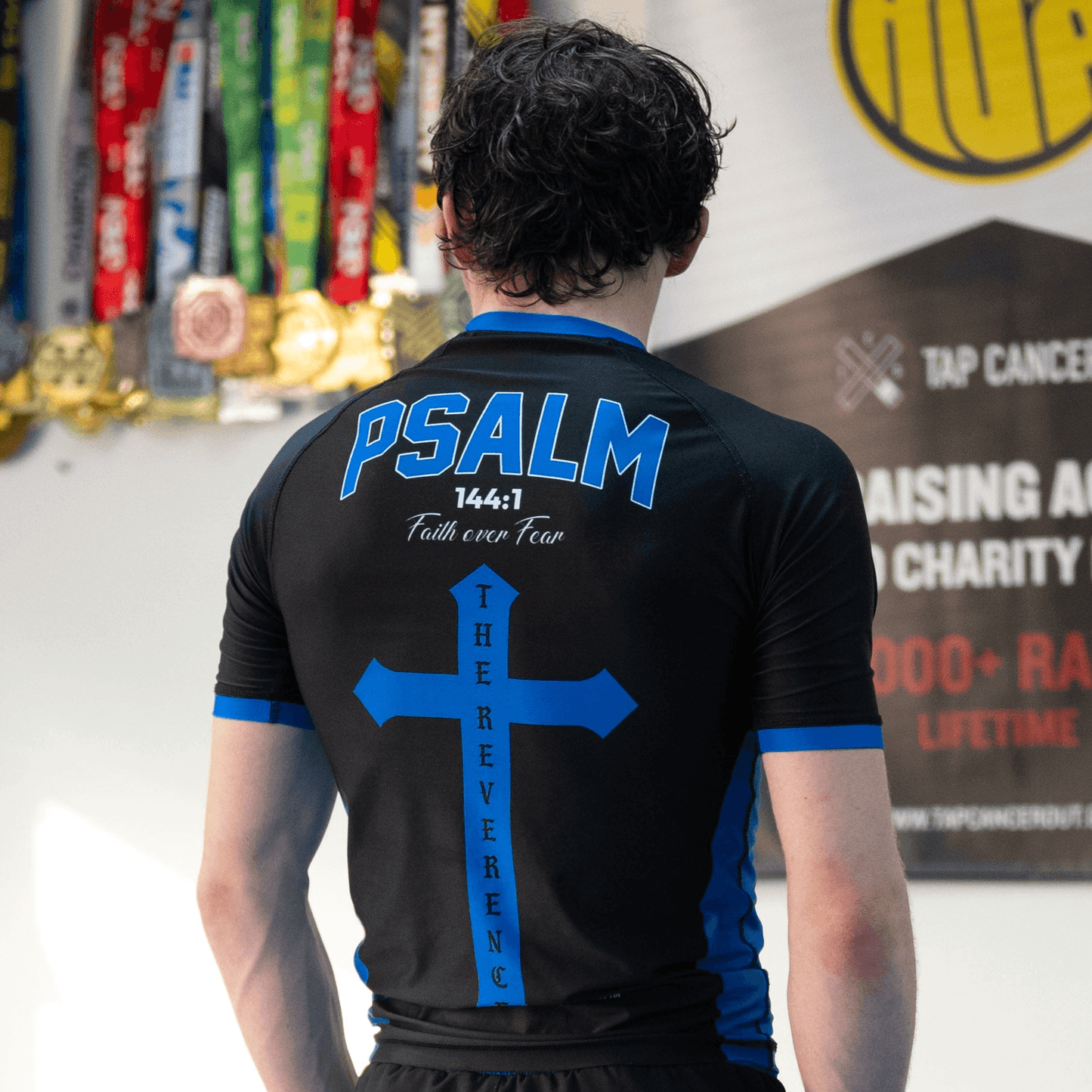 Person wearing a black sports jersey with 'PSALM 144:1' printed on the back, standing in front of a banner.