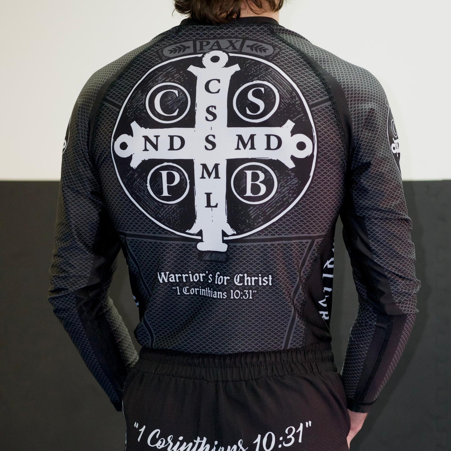 Person wearing a black athletic outfit with a cross design on the back, standing against a white wall.